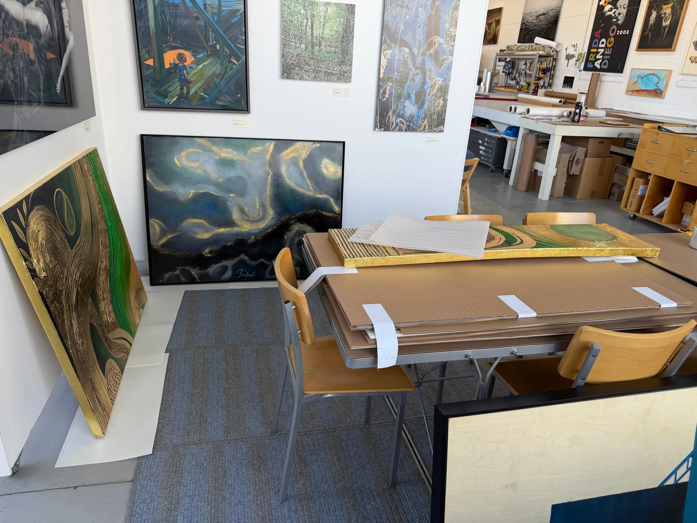 Framed in black and hung in a gallery workshop, beside other paintings and a work table